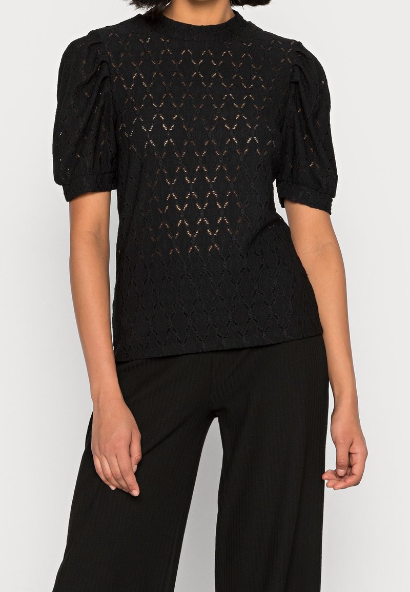 Blouse noire en dentelle à manches courtes bouffantes, portée avec un pantalon noir ample sur une personne, sur un fond clair uni.