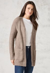 Gilet ouvert beige clair en matériau duveteux, avec des bords côtelés et deux poches avant. Porté sur un t-shirt blanc.