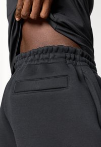 Calças de fato pretas feitas de um tecido macio, com uma cintura elástica e um pequeno bolso traseiro, parcialmente visível por uma parte superior levantada.