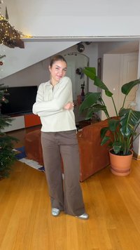 Ανοιχτό πράσινο μακρυμάνικο τοπ με ραβδώσεις, καφέ παντελόνι φαρδύ στο πόδι και ασημί παπούτσια. Εσωτερικός χώρος με φυτά, τηλεόραση και ξύλινο πάτωμα.