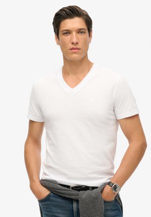 Giovane uomo con capelli corti e scuri, indossa una maglietta bianca a scollatura a V, jeans blu, un maglione grigio annodato in vita e un orologio da polso nero.