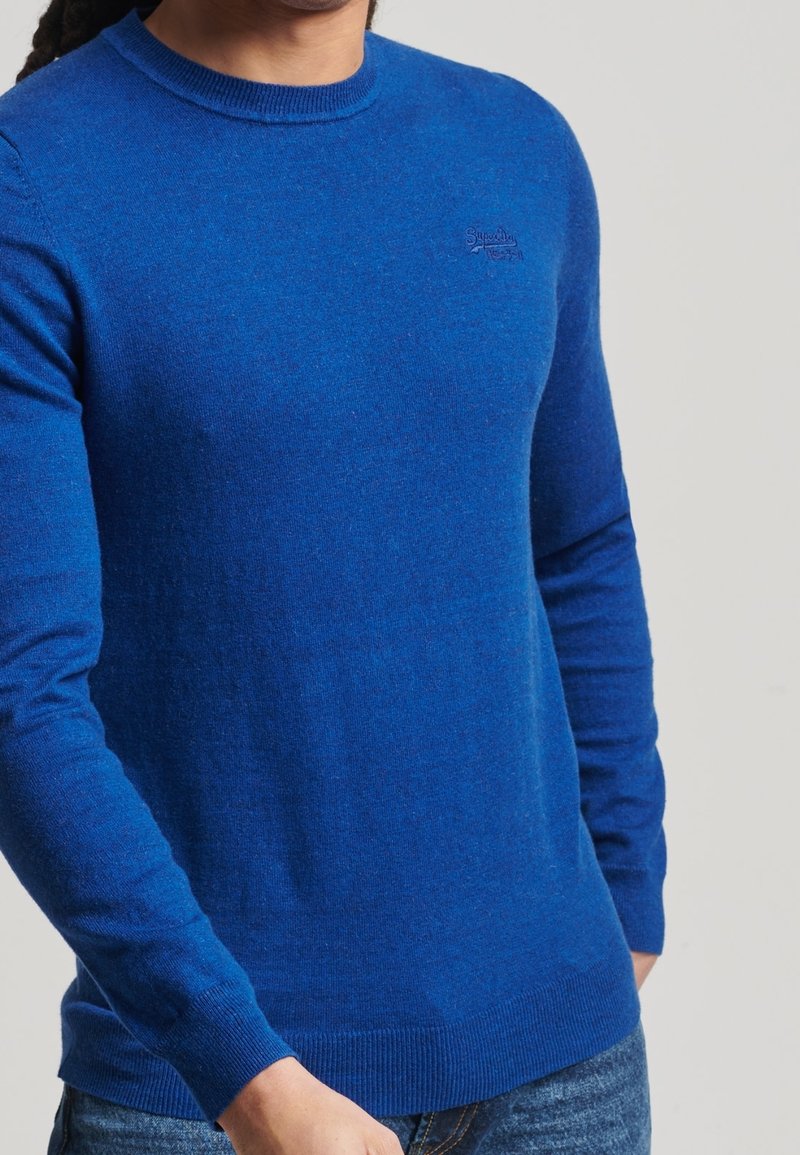 Homme portant un pull bleu vif à manches longues avec poignets et ourlet côtelés, associé à un jean bleu sur fond uni.