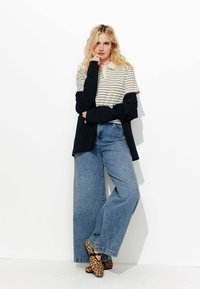 Polo rayé beige et marine, cardigan noir superposé, jeans larges bleus, et ballerines imprimées léopard. Tenue simple et moderne.