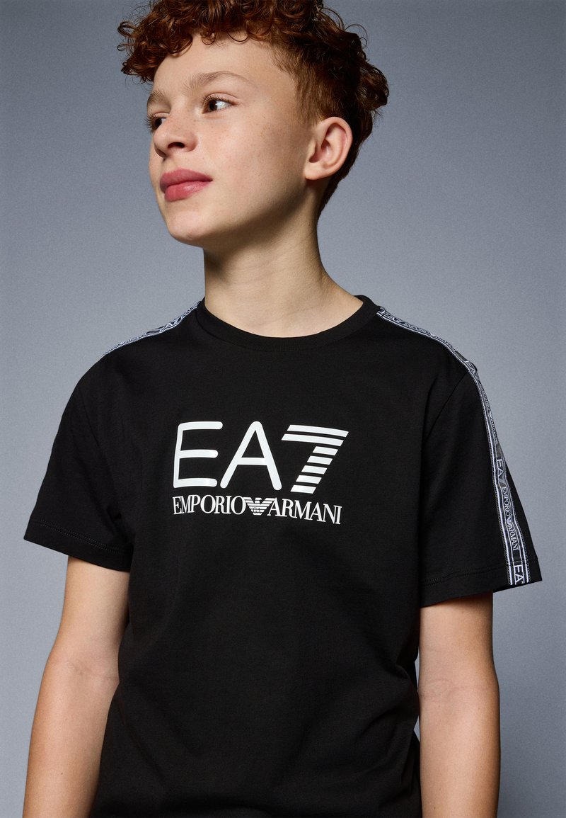 Melnas īsām piedurknēm T-krekls ar balto izdrukāto logo "EA7 Emporio Armani". Sānos ir kontrastējoši rakstaini detaļas. Kokvilnas materiāls.