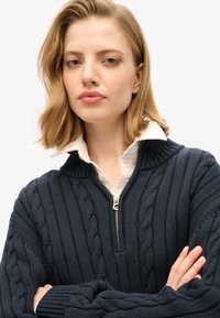 Granatowy sweter na drutach z wzorem warkocza, wysokim prążkowanym kołnierzem i srebrnym zamkiem błyskawicznym. Noszony na białej koszuli zapinanej na guziki. Długie rękawy.