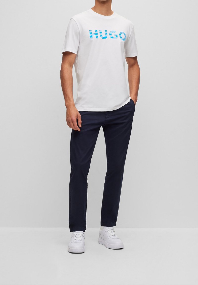 T-shirt en coton blanc avec le logo bleu "HUGO", associé à un pantalon marine et des baskets blanches. Design simple, coupe droite avec des manches courtes.