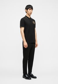 Άνδρας που στέκεται μπροστά σε λευκό φόντο, φοράει μαύρο πόλο μπλουζάκι με χρυσή επένδυση και έμβλημα, μαύρο παντελόνι και μαύρα επίσημα παπούτσια.