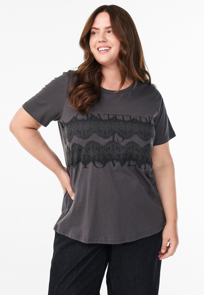 Graues Baumwoll-T-Shirt mit einem schwarzen Spitze-Muster und dem Text "DANKBARKEIT IST KRAFT" in Schwarz. Kurze Ärmel und eine lockere Passform.