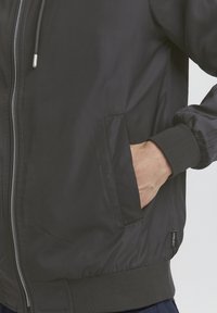 Ruka v kapse černé zipové bundy s patentovými manžetami a lemem, která má poblíž kapsy malou látkovou značku.