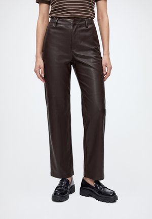 Person iført mørkebrune læder bukser med lige ben, sorte chunky loafers og en brun stribet kortærmet top, der står mod en ensfarvet baggrund.