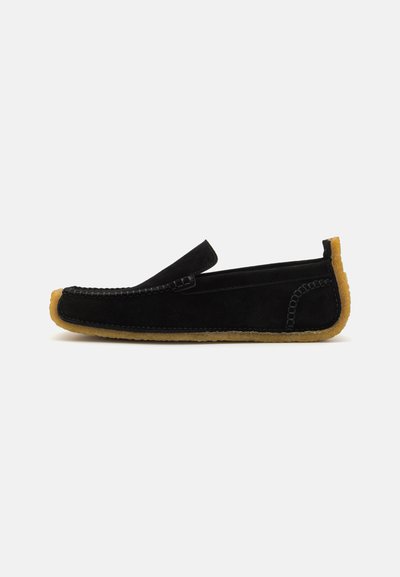 Zwarte suède moccasin-stijl pantoffel met een beige rubberen zool, met stiksels en een instapontwerp. Platte profiel.