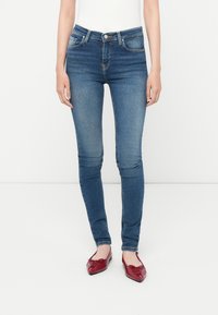 Jeans ajustados de color azul con una cintura de altura media, cinco bolsillos y un ligero desvanecimiento. Se llevan con zapatos planos rojos, mostrando una silueta elegante.
