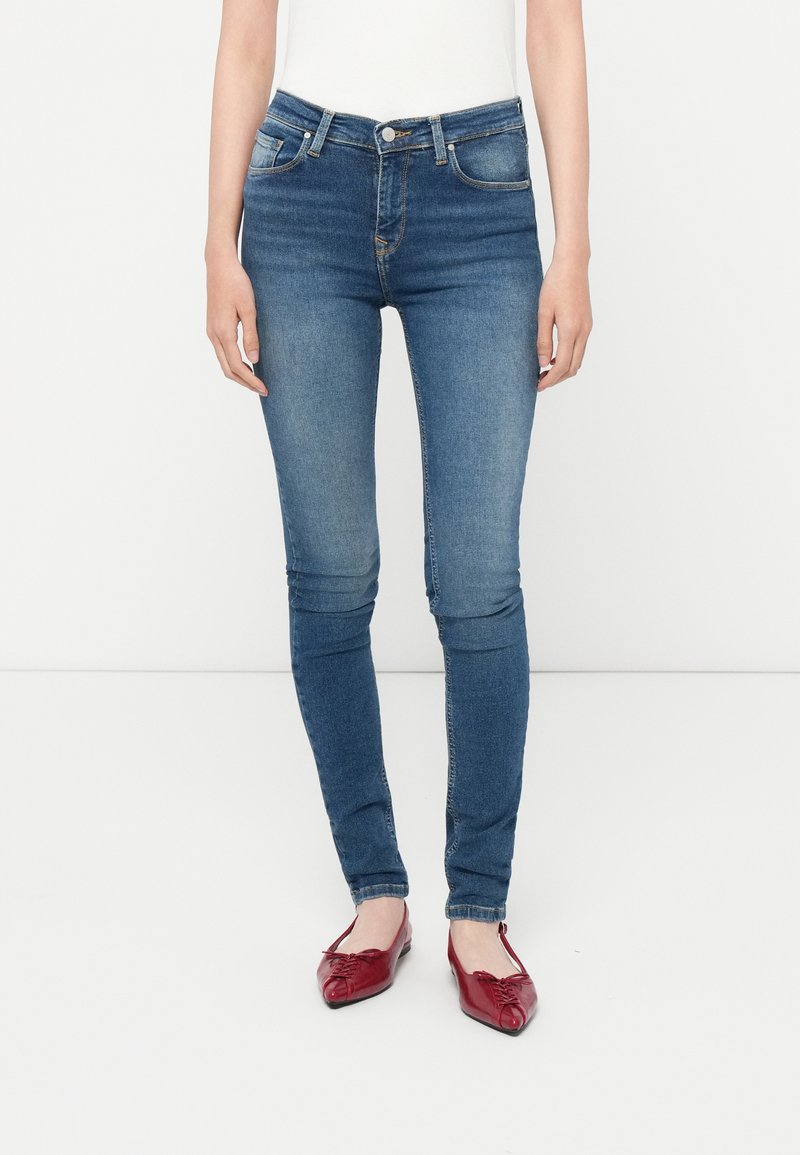 Jeans ajustados de color azul con una cintura de altura media, cinco bolsillos y un ligero desvanecimiento. Se llevan con zapatos planos rojos, mostrando una silueta elegante.
