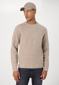 Beige knitted sweater with ribbed texture and round neckline, paired with a light beige cap. The model wears dark jeans.