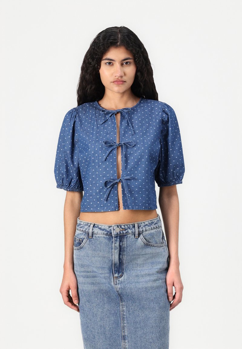 Denimblus med puffärmar, prickigt mönster, nyckelhålsfront med rosetter och kort längd. Matchad med en högmidjad denimkjol.