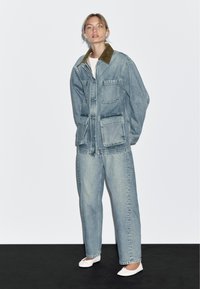 Vrouw staat tegen een witte muur, draagt een oversized spijkerjack met een bruine kraag, bijpassende wijde jeans, een witte top en witte balletschoenen.