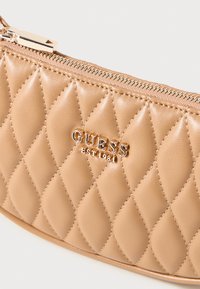 Borsa a tracolla sintetica beige trapuntata con chiusura a zip, caratterizzata da un logo dorato e dettagli di cucitura. Texture liscia con una forma arrotondata.