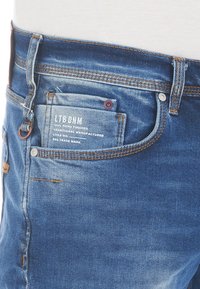 Vaqueros de denim azul con un ajuste regular, con costuras de cobre, un pequeño bolsillo frontal y una etiqueta en la parte trasera. Desvanecimiento sutil en la tela.