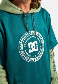 Turkusowa bluza z kapturem z zielonym wnętrzem kaptura. Posiada duży biały nadruk logo oraz czarne sznurki z białymi akcentami na rękawach.