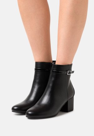 Bottines en cuir noir à talons carrés et petites brides à boucle portées sur des jambes nues sur fond blanc.