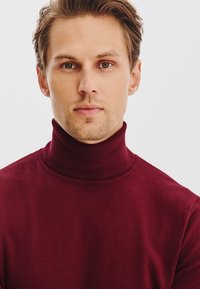 Burgundowy sweter z golfem w miękkim materiale, dopasowany krój z wysokim kołnierzem, minimalistyczna tekstura, noszony przez mężczyznę z krótkimi włosami i lekkim zarostem.