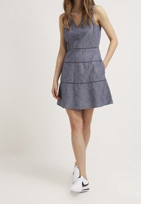 Vestido de denim en forma de A con escote en V, que presenta un diseño en capas y costuras horizontales. Combinado con zapatillas blancas. Textura ligera.