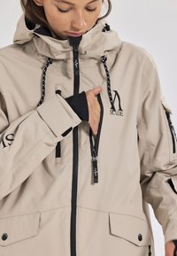 Beige wasserdichte Jacke mit hohem Kragen, verstellbaren Zugbändern, mehreren Reißverschlusstaschen und kontrastierenden schwarzen Akzenten.