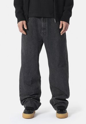 Osoba ubrana w luźne ciemnoszare dżinsy i czarne trampki z gumowymi podeszwami, stojąca na jednolitym białym tle.