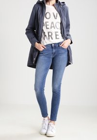 Mörkblå lättviktsjacka med huva över en krämfärgad t-shirt med fet svart text, tillsammans med blå skinny jeans och vita sneakers.