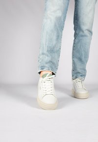 Weiße Ledersneaker mit strukturierter Oberfläche, runder Zehenpartie und flachen Schnürsenkeln, kombiniert mit hellblauen Jeans.