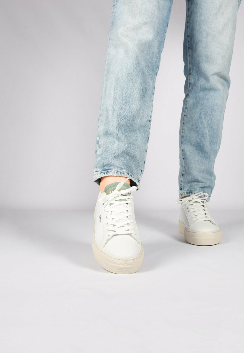 Weiße Ledersneaker mit strukturierter Oberfläche, runder Zehenpartie und flachen Schnürsenkeln, kombiniert mit hellblauen Jeans.