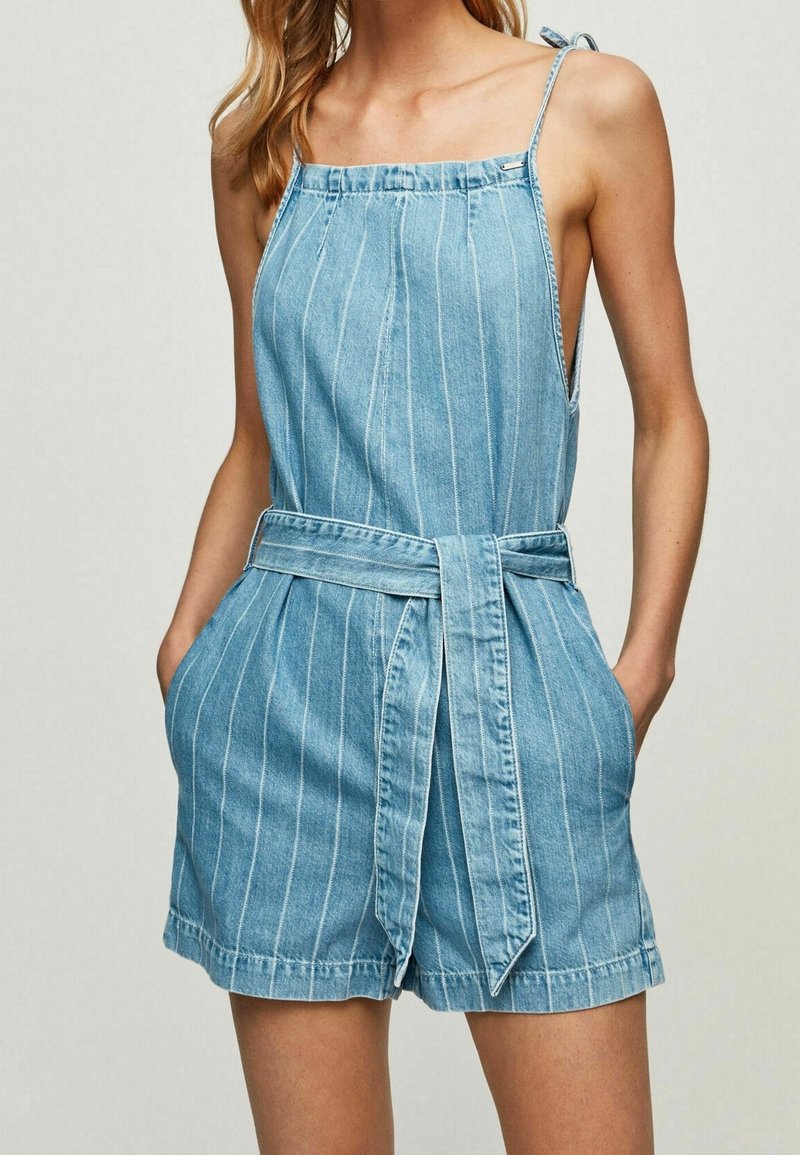 Žena v světle modrém pruhovaném denimu s tenkými ramínky, bočními kapsami a odpovídajícím páskem z látky uvázaným v pase.