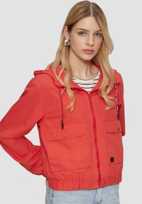 Rote Kapuzenjacke mit zwei Fronttaschen, elastischem Saum und Kordelzug. Getragen über einem gestreiften Hemd mit glatter Textur.
