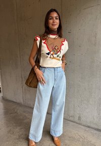 Γυναίκα που στέκεται μπροστά σε τσιμεντένιο τοίχο φορώντας αμάνικο πουλόβερ με σχέδιο καβαλάρη αλόγου, ανοιχτό μπλε τζιν με φαρδιά μπατζάκια, καφέ τσάντα και παπούτσια σε ταμπ χρώμα.