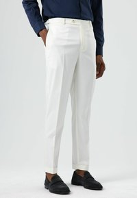 Pantalon blanc cintré, taille ajustée, devant plissé et ourlets retroussés, associé à des mocassins en daim foncé et une chemise bleu marine.