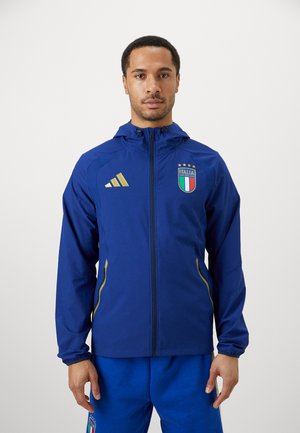 Blå atletisk jakke med hætte, guld accenter og lynlås, med "ITALIA" logo og striber på skuldrene; parret med blå bukser.