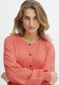 Cardigan corail en coton doux, avec un col rond, des poignets côtelés et des boutons marron sur le devant.
