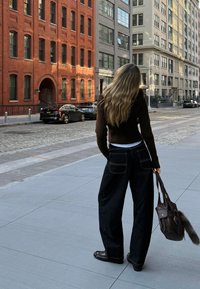 Crne široke traperice s kontrastnim šavovima, smeđi rebrasti pulover i smeđa torba s krznom. Kamenita ulica i ciglene zgrade.