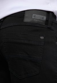 Czarne dżinsy z denimu z skórzaną metką Mustang, kontrastowymi szwami i charakterystycznym wzorem tylnej kieszeni z przeszyciem w kształcie gór.