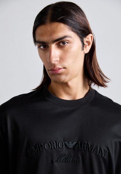 Tânăr cu păr lung și închis la culoare, purtând un hanorac negru Emporio Armani Milano, privind ușor spre o parte pe un fundal simplu.