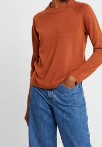 Maglione color ruggine con collo alto, maniche raglan e una consistenza morbida, abbinato a jeans blu chiaro a vestibilità rilassata.