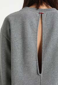 Sudadera gris con detalle de abertura en la parte posterior, textura suave y escote redondo. El diseño incluye costuras en contraste y una pequeña etiqueta.