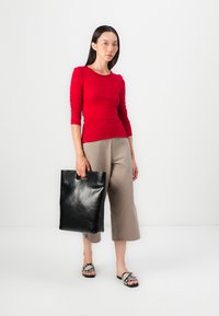 Red fitted long-sleeve top, beige wide-leg pants, black leather tote with cut-out handle, and patterned flat sandals. Smooth textures and vibrant color.