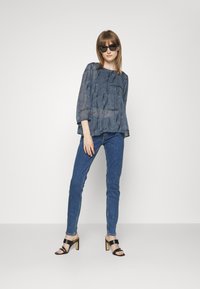 Blusa de mangas compridas, azul com padrão, de corte solto, combinada com jeans de denim justos e sandálias de salto alto pretas. Óculos de sol pretos finalizam o visual.