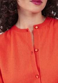 Oranger gestrickter Cardigan mit rundem Ausschnitt, zwei Knöpfen und feiner Textur, teilweise im Bild zu sehen.