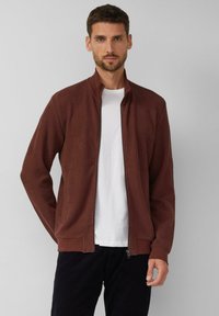 Braune Jacke mit Reißverschluss und gerippter Struktur, Stehkragen und Seitentaschen, getragen über einem weißen T-Shirt und kombiniert mit dunkler Hose.