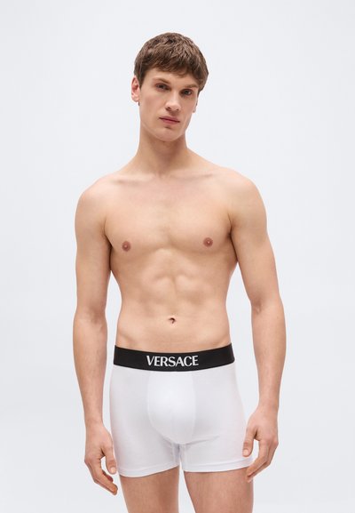 Άνδρας νεαρής ηλικίας χωρίς πουκάμισο, φοράει λευκά μπόξερ με μαύρη ζώνη μέσης με ετικέτα "VERSACE", στέκεται μπροστά σε απλό φωτεινό φόντο.