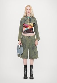 Giacca di jeans verde con design cropped, indossata sopra un top tie-dye multicolore. Abbinata a pantaloni corti di lunghezza al ginocchio, stivali neri alla caviglia e una borsa con frange.