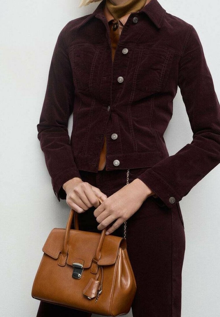 Personne portant une veste boutonnée marron foncé et un pantalon, tenant un sac à main en cuir fauve de taille moyenne avec une fermeture métallique.