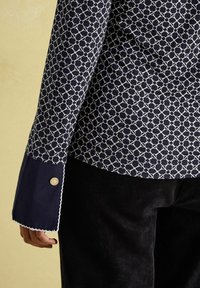 Navyblaues Hemd mit weißem geometrischen Muster, ausgestattet mit einem kontrastierenden navyblauen Manschetten- und Knopfdetail. Weicher Stoff und strukturierte Passform.